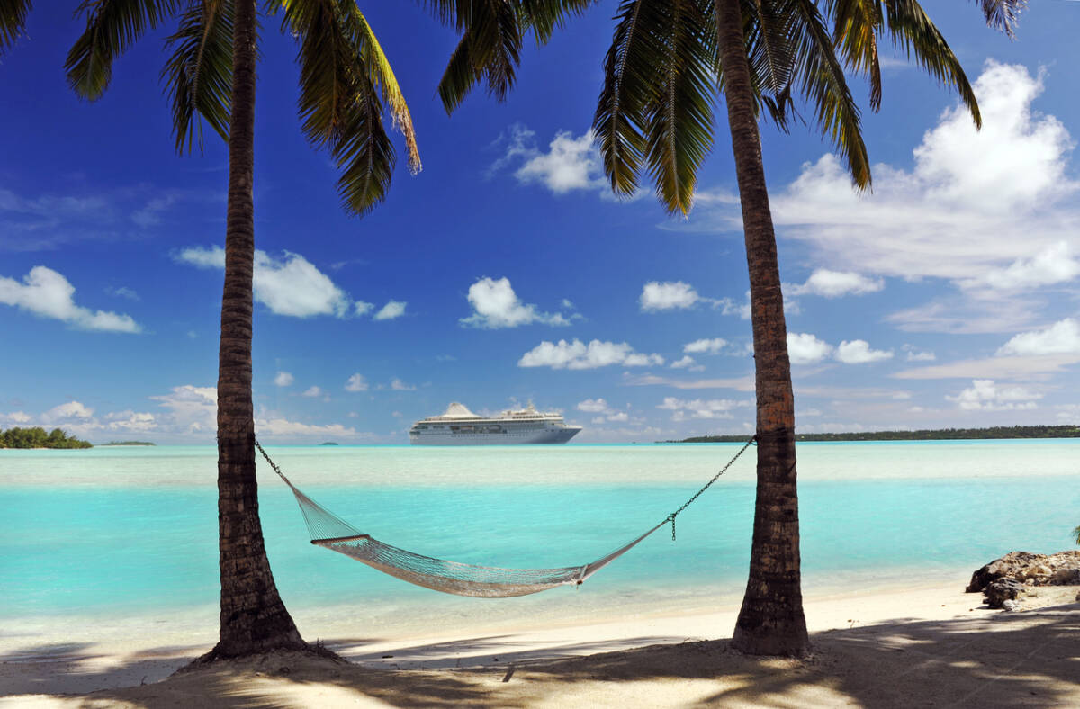 A cruise ship passes an idyllic tropical island in the South Pacific with hammock and palm trees in semi-silhouette